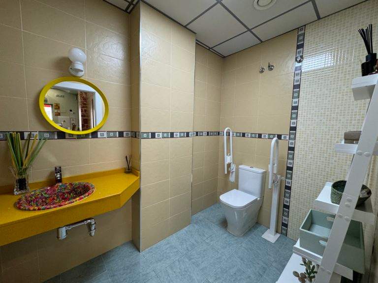 Baño adaptado con barras de apoyo, lavabo amarillo y suelo antideslizante
