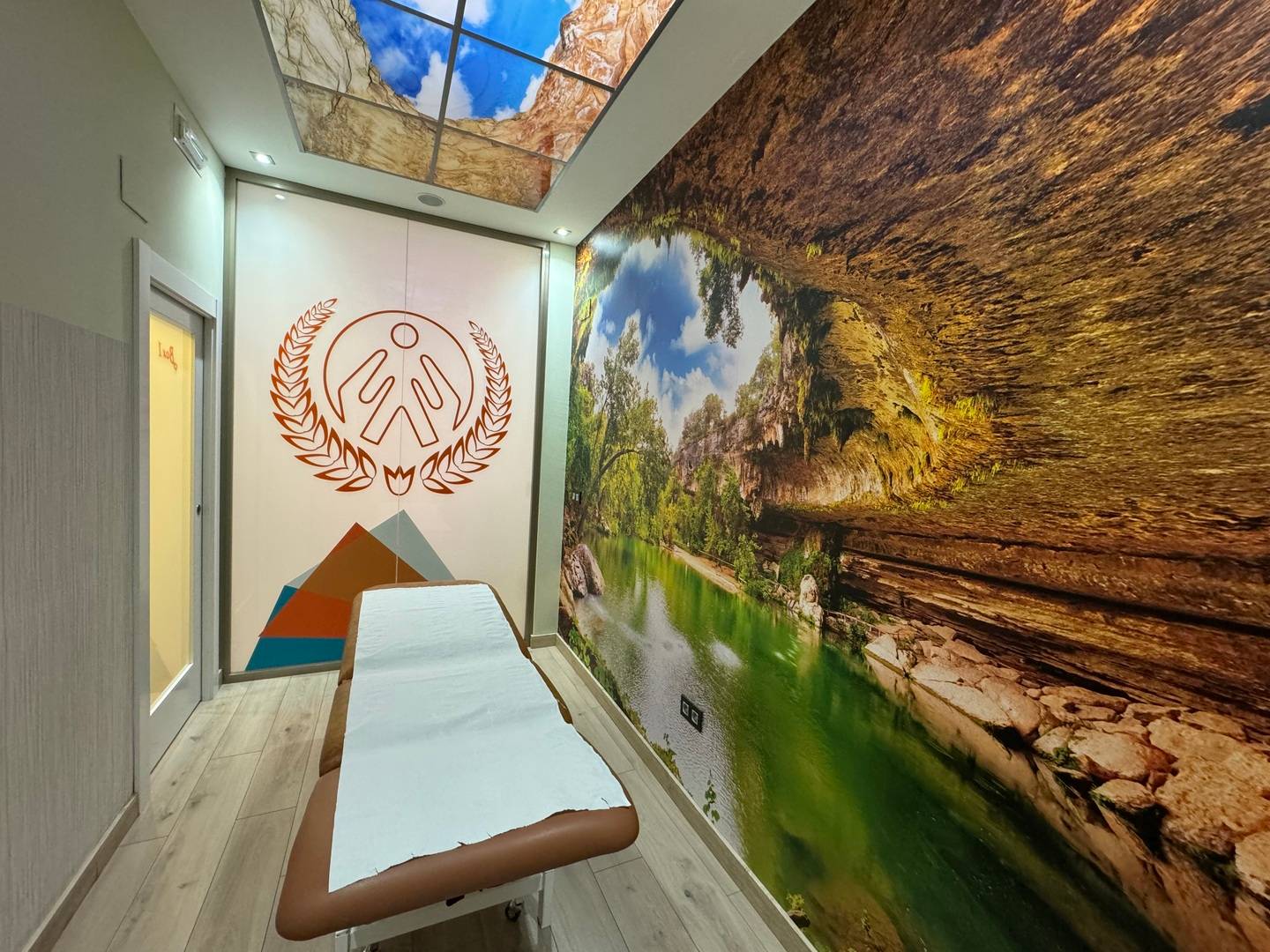 Sala de tratamiento con mural de cueva y lago, techo retroiluminado con cielo abierto